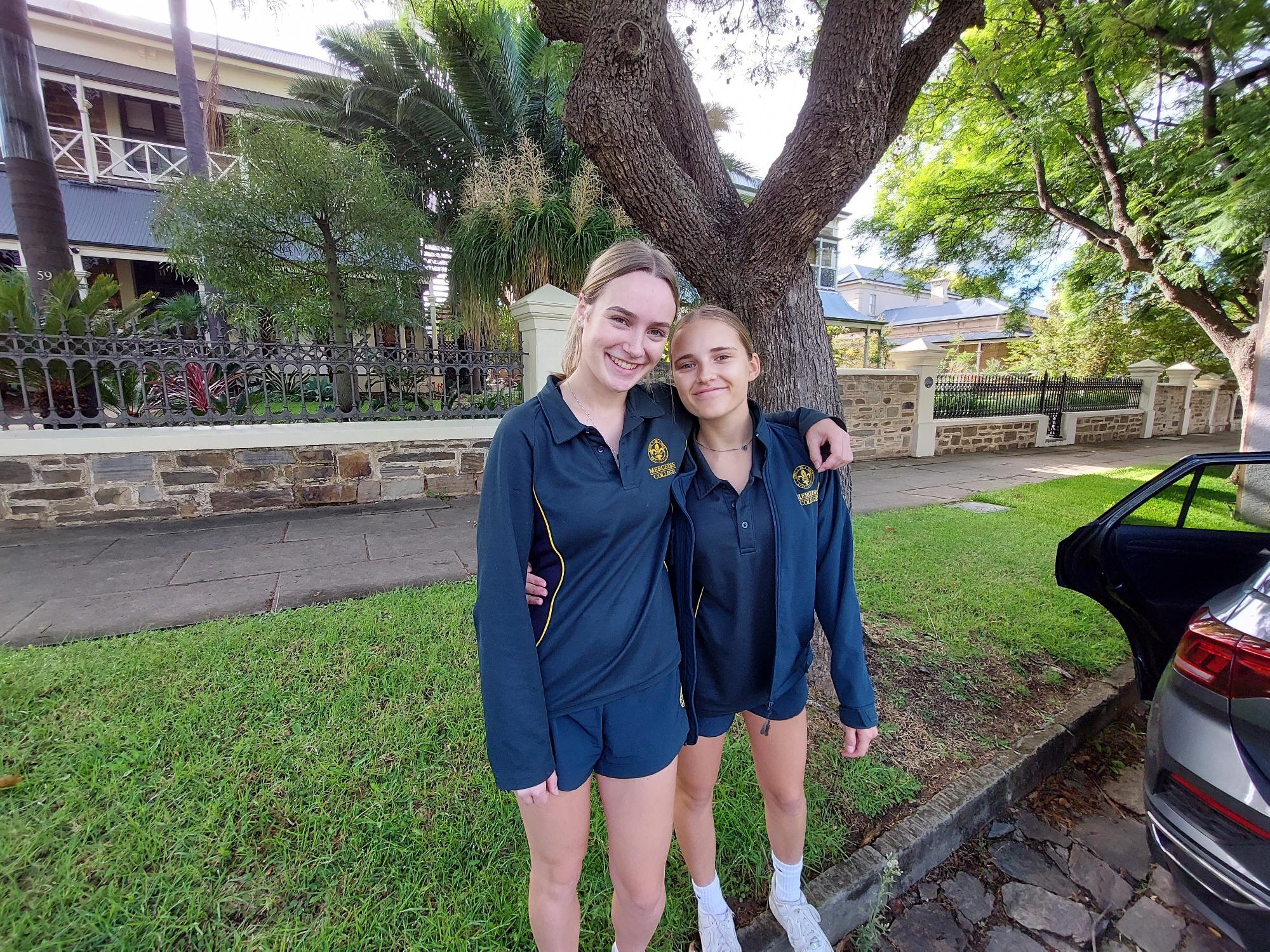 Mercedes College girls standing arm in arm in sports uniforms outdoors near a tree and parked car.